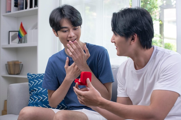 Sorprendido joven levantando las manos y sonriendo mientras su novio le propone matrimonio en la sala de estar Pareja emocional enamorada compromiso Concepto de estilo de vida de pareja LGBT gay asiática