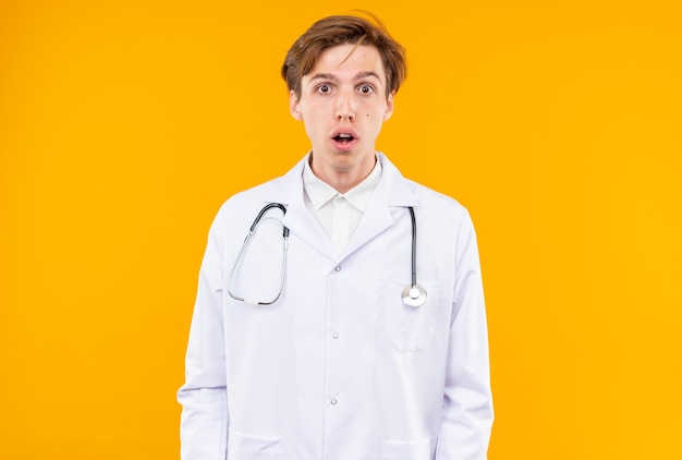 Sorprendido joven médico vistiendo bata médica con estetoscopio aislado en la pared naranja