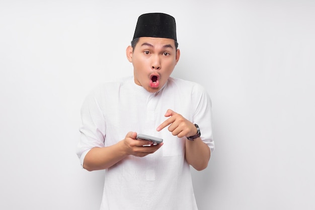 Sorprendido joven musulmán asiático vestido con ropa árabe señalando con el dedo al teléfono inteligente con la boca abierta aislada en el fondo blanco Concepto de estilo de vida islámico religioso de la gente