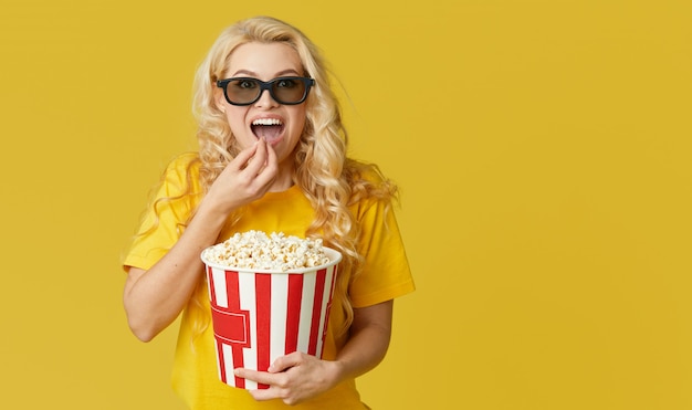 Sorprendido joven rubia en gafas 3d y camisa amarilla comiendo palomitas de maíz, se ve una película impactante en el cine. Aislado