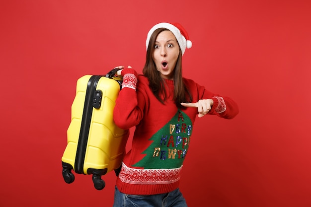 Sorprendido joven Santa manteniendo la boca abierta, apuntando con el dedo índice en la maleta amarilla aislada sobre fondo rojo de la pared. Feliz año nuevo 2019 celebración concepto de fiesta navideña. Simulacros de espacio de copia.