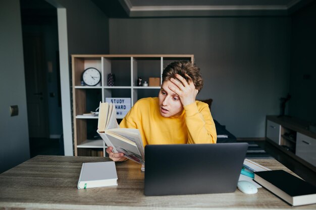 Sorprendido joven sentado en una mesa en una habitación en casa leyendo un libro y disfrutando de Internet Retrato de un estudiante emotivo que estudia remotamente en casa Cuarentena