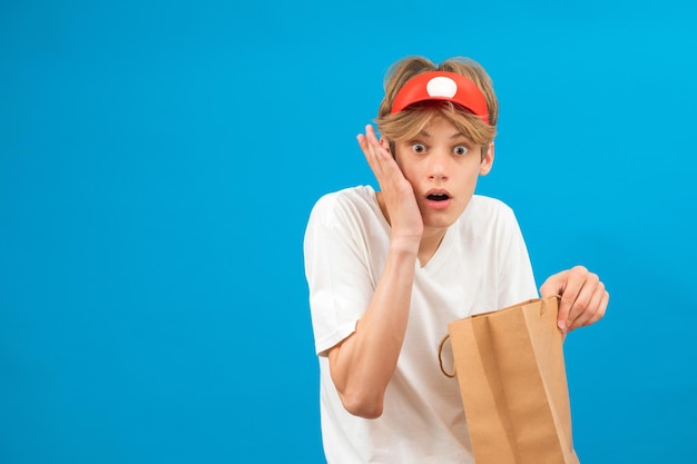 Sorprendido joven vendedor en camiseta blanca casual aislada sobre fondo azul retrato de estudio Concepto de estilo de vida de la gente Maqueta espacio de copia Adolescente sosteniendo bolsa de paquete con compras