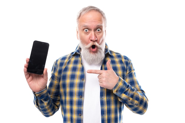 Sorprendido jubilado senior hombre canoso con barba en una camisa sosteniendo un teléfono inteligente en un blanco