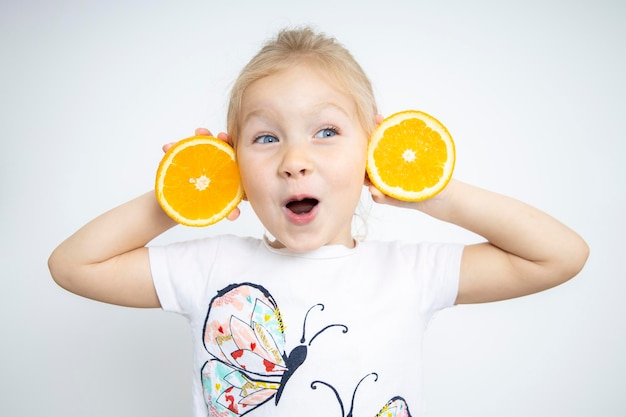 Sorprendido niño niña rubia sosteniendo dos mitades cortadas de una naranja sobre un fondo blanco.