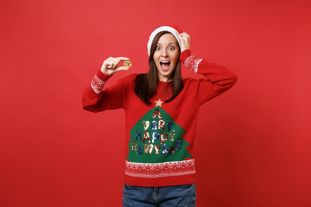 Sorprendido Santa niña poniendo la mano en la cabeza sosteniendo bitcoin moneda futura moneda de metal de color dorado aislado sobre fondo rojo. Feliz año nuevo 2019 celebración concepto de fiesta navideña. Simulacros de espacio de copia.