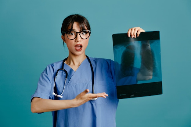 Sorprendido sosteniendo y apuntando a la joven doctora de rayos x con estetoscopio uniforme aislado en el fondo azul