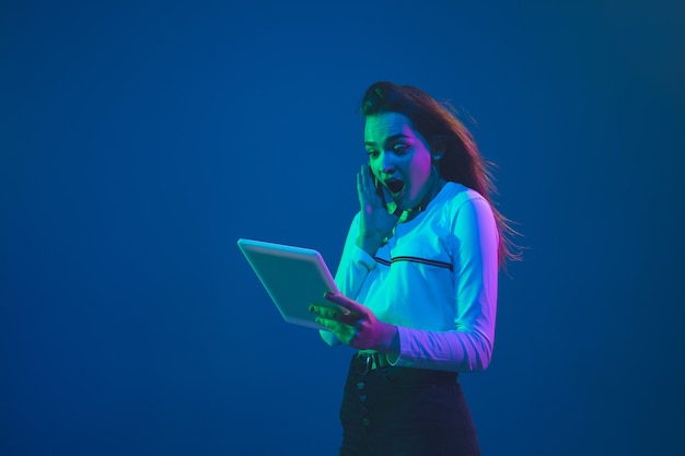 Sorprendido con tableta. Retrato de mujer joven caucásica aislado sobre fondo azul de estudio en luz de neón. Modelo de mujer hermosa. Concepto de emociones humanas, expresión facial, ventas, publicidad, cultura juvenil.