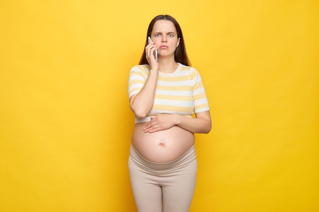 Sorpresa conmocionada mujer embarazada caucásica con el vientre desnudo usando una camiseta casual aislada sobre un fondo amarillo de pie con un teléfono inteligente hablando por teléfono móvil llamando al médico