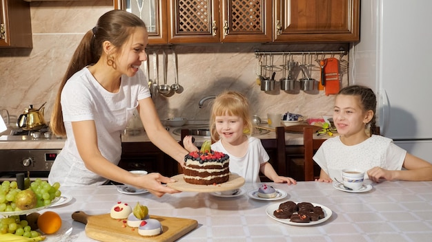 Sorpresa de cumpleaños de mamá. La madre trae el pastel casero de chocolate a sus hijas tomando un té sentado en la cocina en casa.