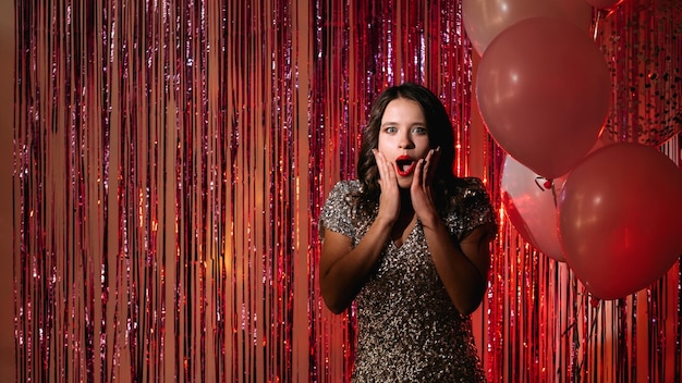 Sorpresa de cumpleaños Mujer asombrada Situación inesperada Emocionada hermosa dama elegante vestido de lentejuelas que parece sorprendida en rojo brillante cascada cortina globos de luz decoración espacio de copia