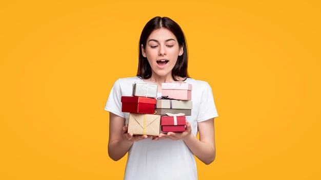 Sorpresa de cumpleaños Regalo de vacaciones Mujer sorprendida con la boca abierta en camiseta blanca sosteniendo pilas de cajas de regalo aisladas en el espacio de copia naranja Día especial Saludo felicitación