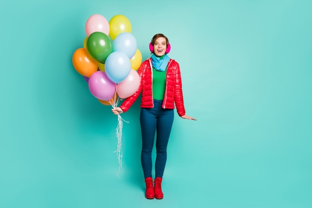 ¡Sorpresa! Foto de cuerpo entero de la fiesta de cumpleaños de la dama emocionada traer globos de aire coloridos usar bufanda de abrigo rojo casual orejeras rosadas pantalones de puente zapatos aislados pared de color verde azulado
