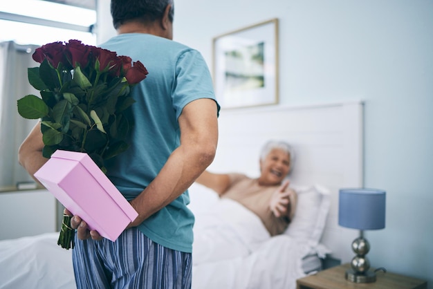 Sorpresa de regalo y una pareja mayor en su aniversario en el dormitorio de su casa juntos para celebrar en la mañana Caja de cumpleaños de flores y un anciano que le da un regalo a su esposa en casa