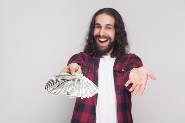 ¡Sorpresa para ti! Retrato de joven empresario afortunado en camisa a cuadros roja y pelo largo y rizado negro de pie, dando y compartiendo fan de dinero. Tiro de estudio interior, aislado sobre fondo gris.