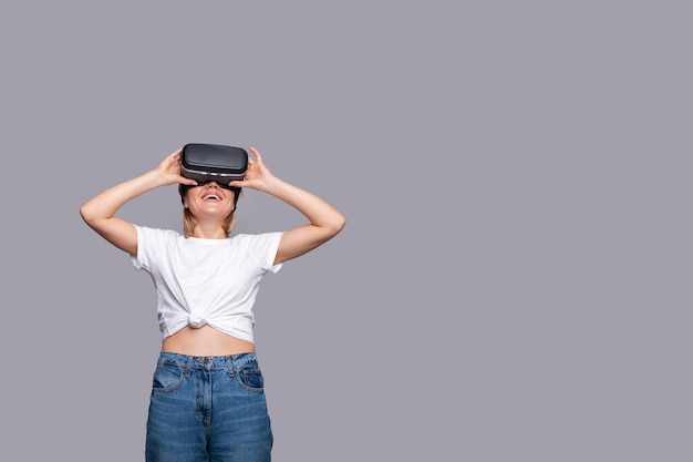 Sorria mulher feliz obtendo experiência usando óculos VRheadset de realidade virtual isolado sobre fundo cinza com espaço de cópia Olhando para cima