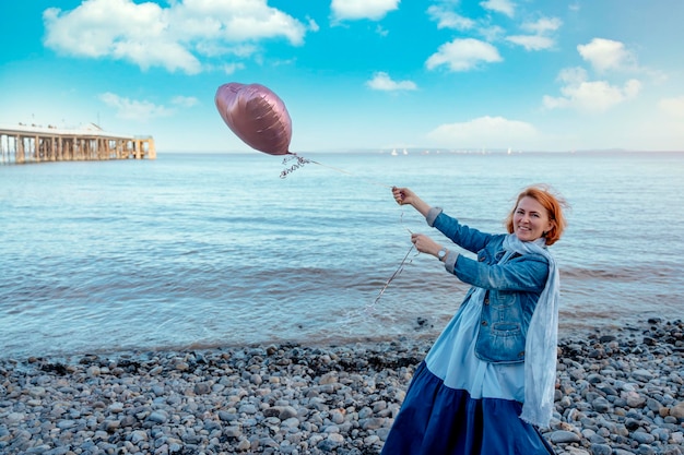 Sorria mulher vermelha de vestido azul e óculos segurando um balão à beira-mar ao pôr do sol