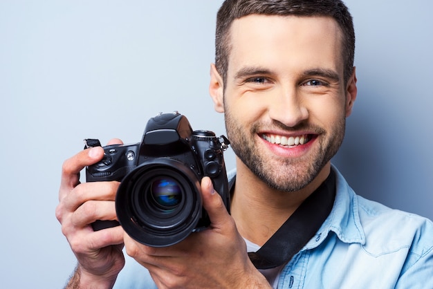 Sorria para a câmera! Jovem bonito segurando uma câmera digital e sorrindo em pé contra um fundo cinza