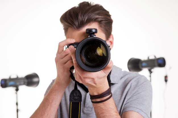 Sorria para a câmera! Jovem com uma camisa pólo fotografando você com uma câmera digital enquanto está no estúdio com equipamento de iluminação no fundo