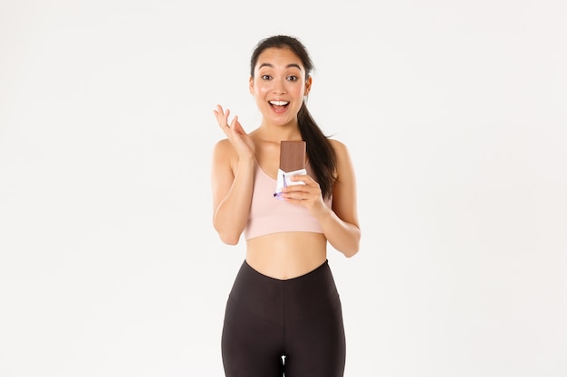 Sorridente alegre garota asiática magro fitness, mulher em roupas esportivas segurando uma barra de chocolate, parecendo despreocupada, refeição de trapaça após um bom treino.