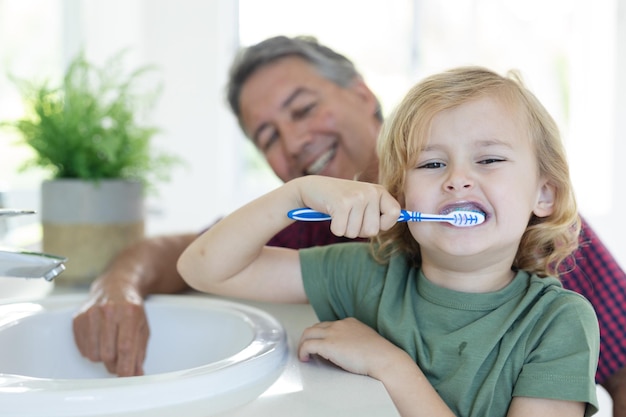 Sorridente avô caucasiano no banheiro ajoelhado ao lado do neto escovando os dentes olhando para a câmera