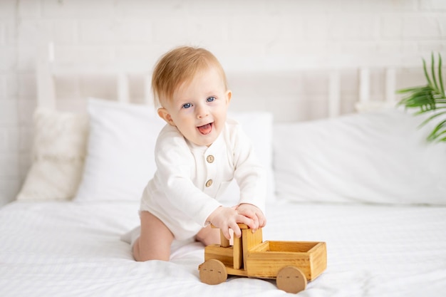 Sorridente bebê loiro de 6 meses de idade está sentado em uma cama grande em um quarto brilhante e brincando com um carro de brinquedo de madeira em uma roupa de algodão o conceito de artigos infantis