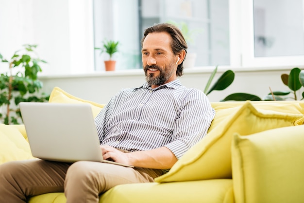 Sorridente, bonito homem barbudo sentado no sofá amarelo e olhando para a tela de um laptop moderno no colo