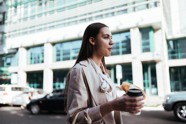 Sorridente e ansioso, a garota se move pela rua da cidade e mastiga uma torta enquanto bebe seu café