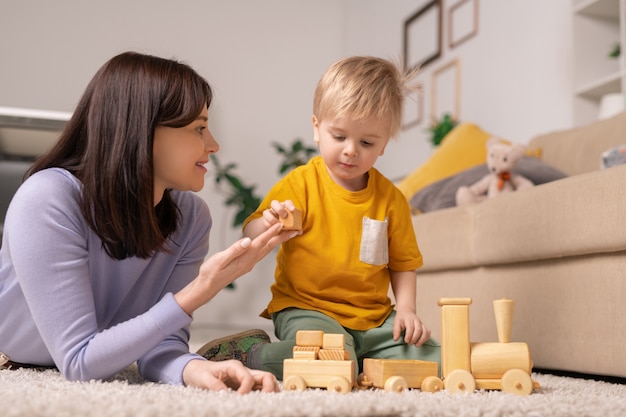 Sorridente e atraente jovem mãe deitada no tapete dando um bloco de madeira ao filho enquanto o ensina a empilhar blocos de brinquedo