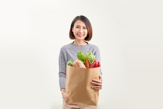 Sorridente e feliz jovem dona de casa asiática segura saco de papel com produtos alimentares frescos em fundo branco