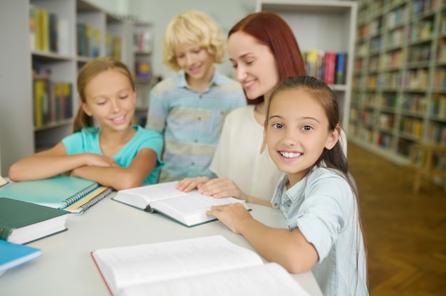 Sorridente e fofa colegial de cabelos escuros sentada à mesa com seus colegas e a professora em uma biblioteca pública