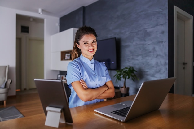 Sorridente e simpática jovem enfermeira sentada em casa com os braços cruzados e usando tecnologias para dar conselhos.