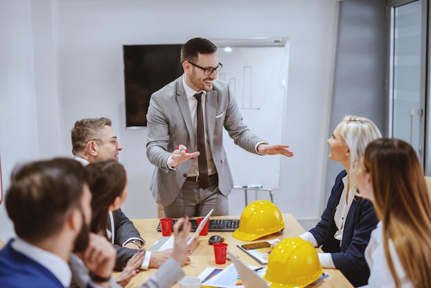 Sorridente empresário bem sucedido em pé e falando sobre o novo projeto enquanto sua equipe sentado na sala de reuniões e fazendo perguntas. Se você deseja alcançar a grandeza, pare de pedir permissão.