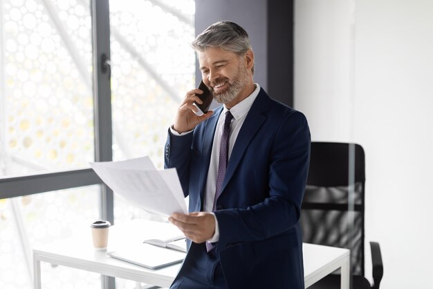 Foto sorridente empresário de meia-idade falando no celular e verificando papéis no escritório