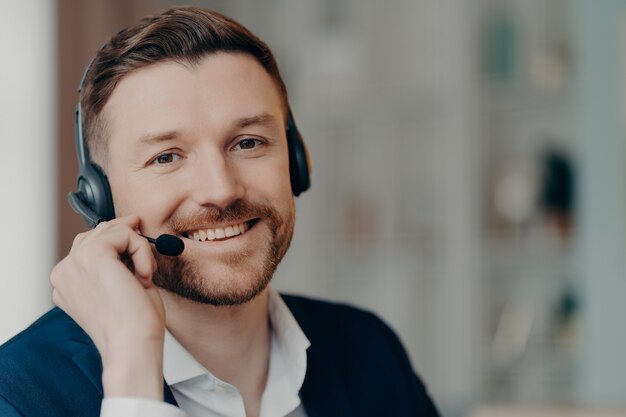 Sorridente empresário habilidoso com barba estando de bom humor usa fone de ouvido vestido em poses de roupas formais contra um fundo desfocado. Oportunidades de trabalho remotas. Comunicação à distância por teleconferência