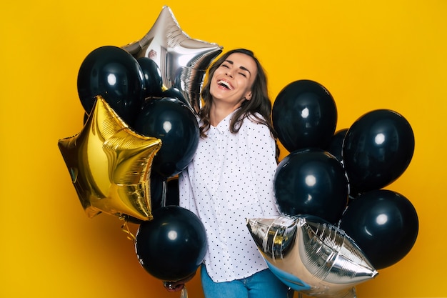 Foto sorridente, feliz e animada linda garota segurando muitos balões pretos nas mãos está posando no fundo amarelo enquanto fazia compras na black friday