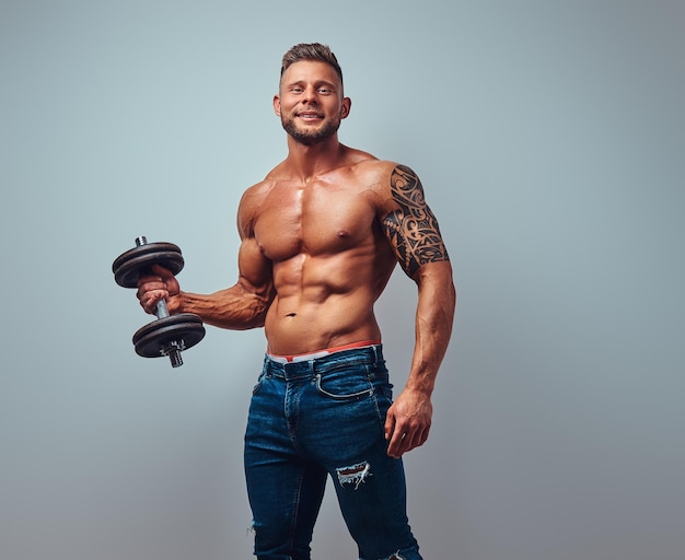 Sorridente fisiculturista sem camisa bonito com corte de cabelo elegante e barba, com tatuagem no braço, posando com halteres em um estúdio. isolado em um fundo cinza.