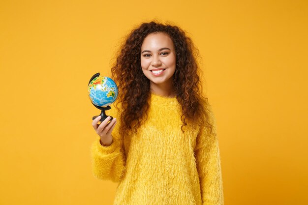 Sorridente jovem afro-americana em suéter de pele posando isolado em fundo amarelo laranja, retrato de estúdio. Conceito de estilo de vida de emoções sinceras de pessoas. Mock up espaço de cópia. Segurando o globo do mundo.