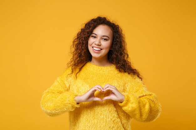 Sorridente jovem afro-americana em suéter de pele posando isolado no fundo da parede amarela laranja. conceito de estilo de vida de pessoas. mock up espaço de cópia. mostrando forma de coração com as mãos, sinal em forma de coração.