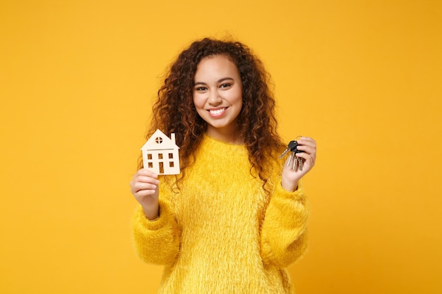 Sorridente jovem afro-americana em suéter de pele posando isolado no fundo da parede laranja amarela no estúdio. Conceito de estilo de vida de pessoas. Mock up espaço de cópia. Segurando em casa de mãos e molho de chaves.