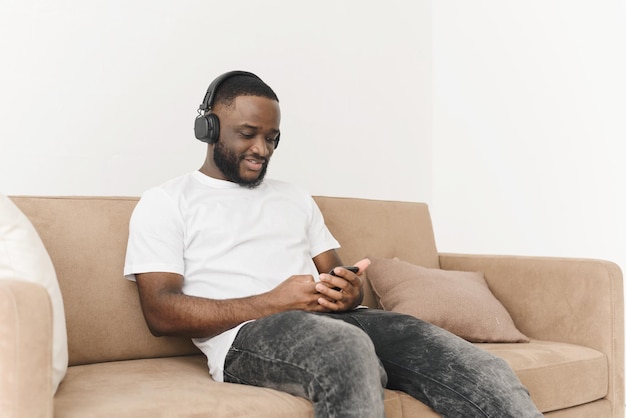 Sorridente jovem afro-americano em fones de ouvido ouvindo música móvel navegando nas redes sociais usando smartphone relaxando enquanto está sentado no sofá em casa