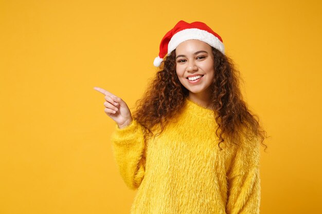 Sorridente jovem afro-americano Papai Noel no suéter de pele, chapéu de Natal isolado em fundo amarelo. Feliz ano novo 2020 conceito de feriado de celebração. Simule o espaço da cópia. Apontando o dedo indicador para o lado.