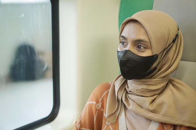 Sorridente jovem asiática em hijab usando máscara sentada em um trem e olhando pela janela