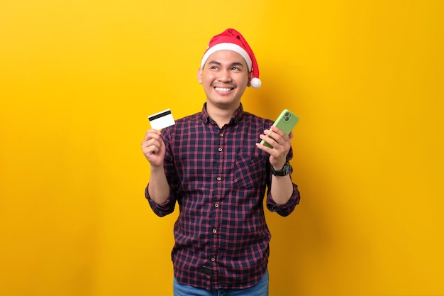 Sorridente jovem asiático com chapéu de Papai Noel usando telefone celular e cartão de crédito olhando para cima sobre o fundo amarelo do estúdio Feliz Ano Novo 2023 celebração feliz conceito de férias