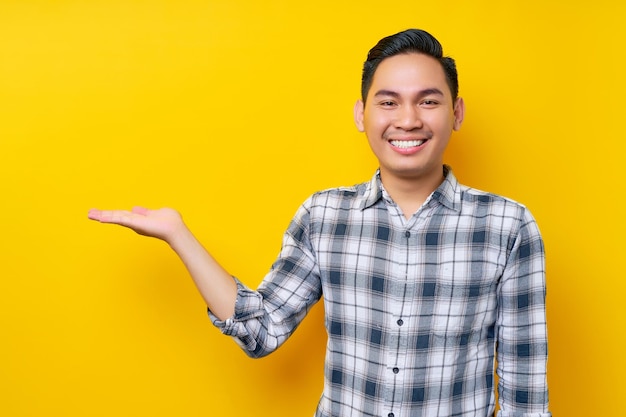 Sorridente jovem asiático em roupas casuais, apresentando espaço de cópia em uma palma da mão isolada em fundo amarelo Conceito de estilo de vida de pessoas