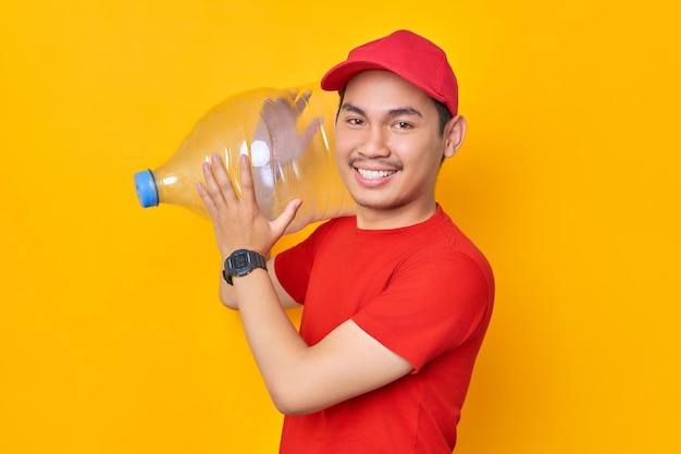 Sorridente jovem asiático em uniforme de camiseta de boné vermelho trabalha como correio de revendedor segurando garrafa de água isolada em fundo amarelo conceito de serviço de entrega profissional