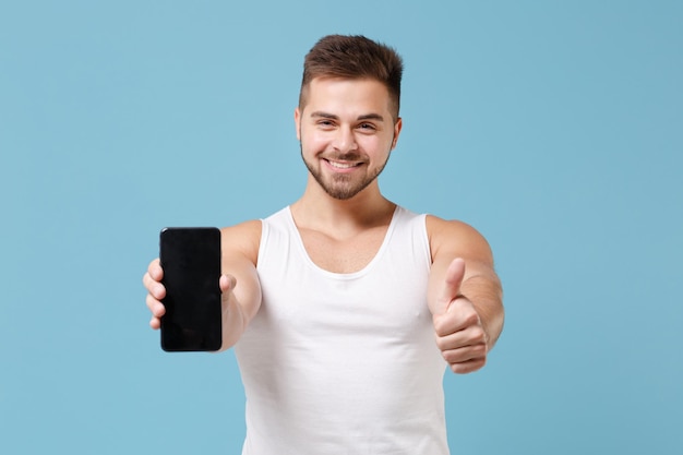 Sorridente jovem barbudo em camiseta branca isolada em fundo azul pastel. Conceito de estilo de vida saudável de fitness esportivo. Mock up espaço de cópia. Segure o celular com tela vazia em branco, aparecendo o polegar.