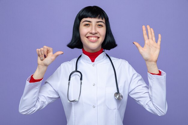 Sorridente, jovem, bonita, caucasiana, com uniforme de médico e estetoscópio fica com a mão levantada e aponta para o lado