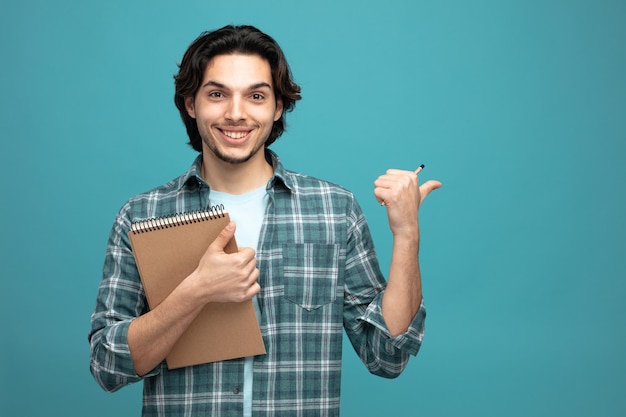 sorridente jovem bonito segurando o bloco de notas e lápis olhando para a câmera apontando para o lado isolado no fundo azul com espaço de cópia