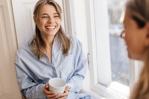 Sorridente jovem caucasiana passa o tempo com um velho amigo sobre a xícara de café, sentado pela janela. Conceito de relaxamento e paz.